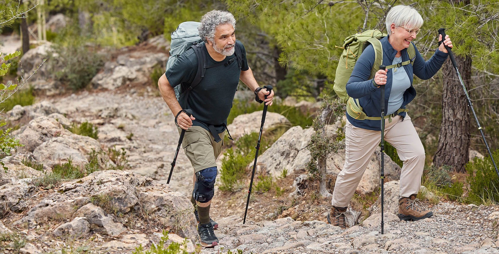 Ein Mann und eine Frau wandern auf einem steinigen Weg durch einen Wald. Beide tragen Wanderausrüstung mit Rucksäcken und Wanderstöcken. Der Mann trägt eine GenuTrain OA Knieorthese am rechten Knie, die seine Bewegung beim Anstieg unterstützt.