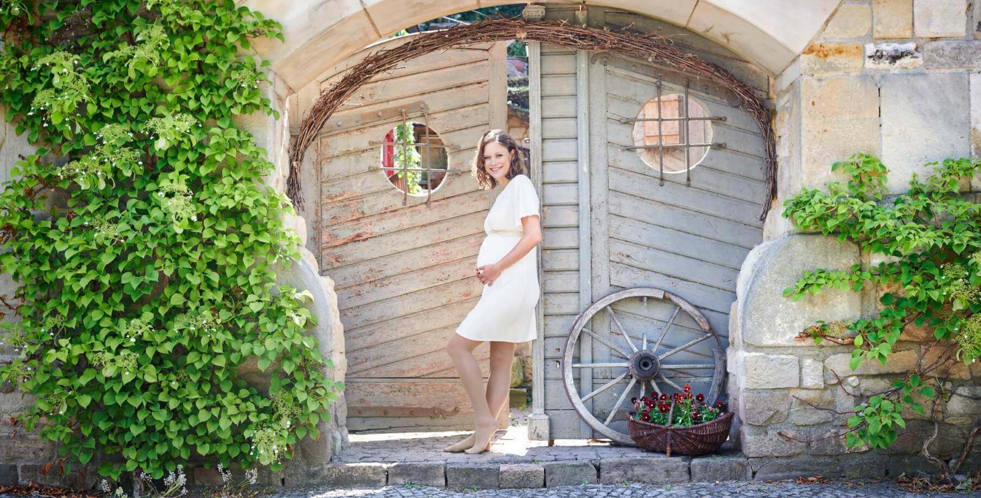 Tipps gegen schwere Beine in der Schwangerschaft Eine schwangere Frau in einem weißem Kleid steht vor einem Gartentor. Sie hat die Hände auf ihren Babybauch abgelegt und lächelt freundlich in die Kamera. Sie trägt VenoTrain micro Kompressionsstrümpfe von Bauerfeind, welche die werdende Mutter optimal versorgt.
