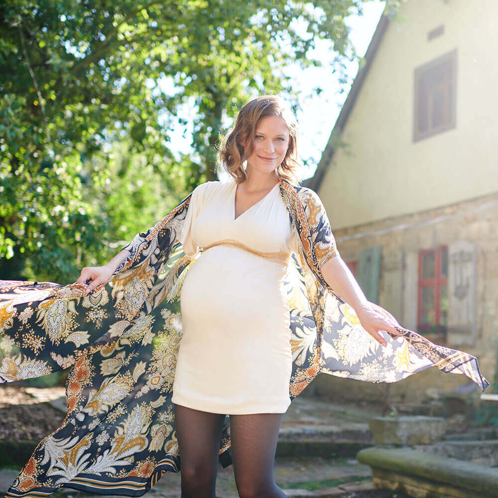 Aktiv die Durchblutung fördern Eine schwangere Frau im weißen Kleid steht in einem Hof und lächelt freundlich in die Kamera. Sie trägt einen Cardigan, welcher im Winde weht. Außerdem hat sie eine VenoTrain® micro Schwangerschaftsstrumpfhose von Bauerfeind an, welche die werdende Mutter optimal versorgt.